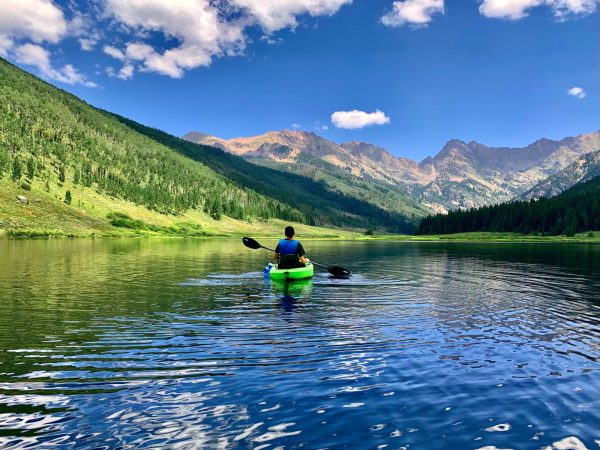 Person in a bright green kayak paddles across a calm alpine lake, with forested green hills and rugged mountains under a blue sky.