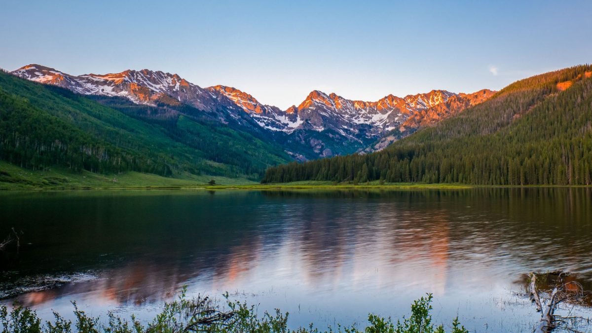 Snow-capped mountains glow orange at sunset behind a green forested valley and calm lake surface. (Alpine landscape)